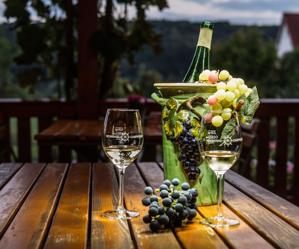 Eine Weinflasche in einem dekorativen Halter mit Weintrauben, zwei gefüllte Weingläser und Weintrauben sitzen auf einem Holztisch im Freien in Neumanns Dampfschiff Gaststätte, mit einem unscharfen Hintergrund aus Grünpflanzen und einem Haus.