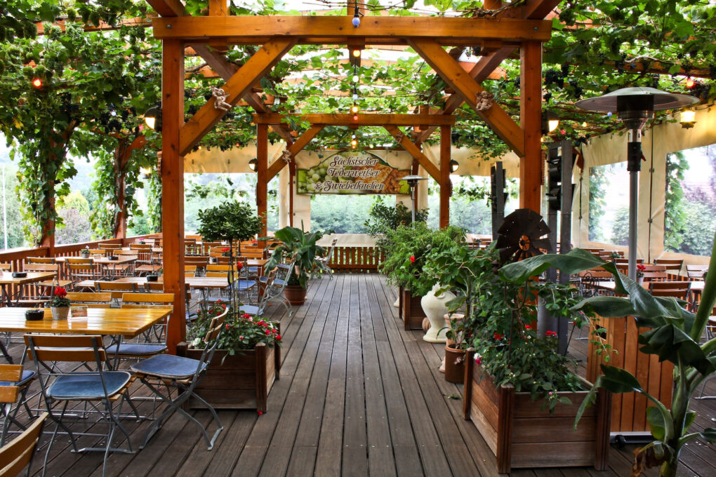 Die hölzerne Außenterrasse des Biergartens Sörnewitz mit Tischen und Stühlen unter einer mit grünen Ranken bewachsenen Pergola, umgeben von Topfpflanzen und kleinen Bäumen, schafft eine gemütliche, einladende Atmosphäre, die zum Entspannen einlädt.