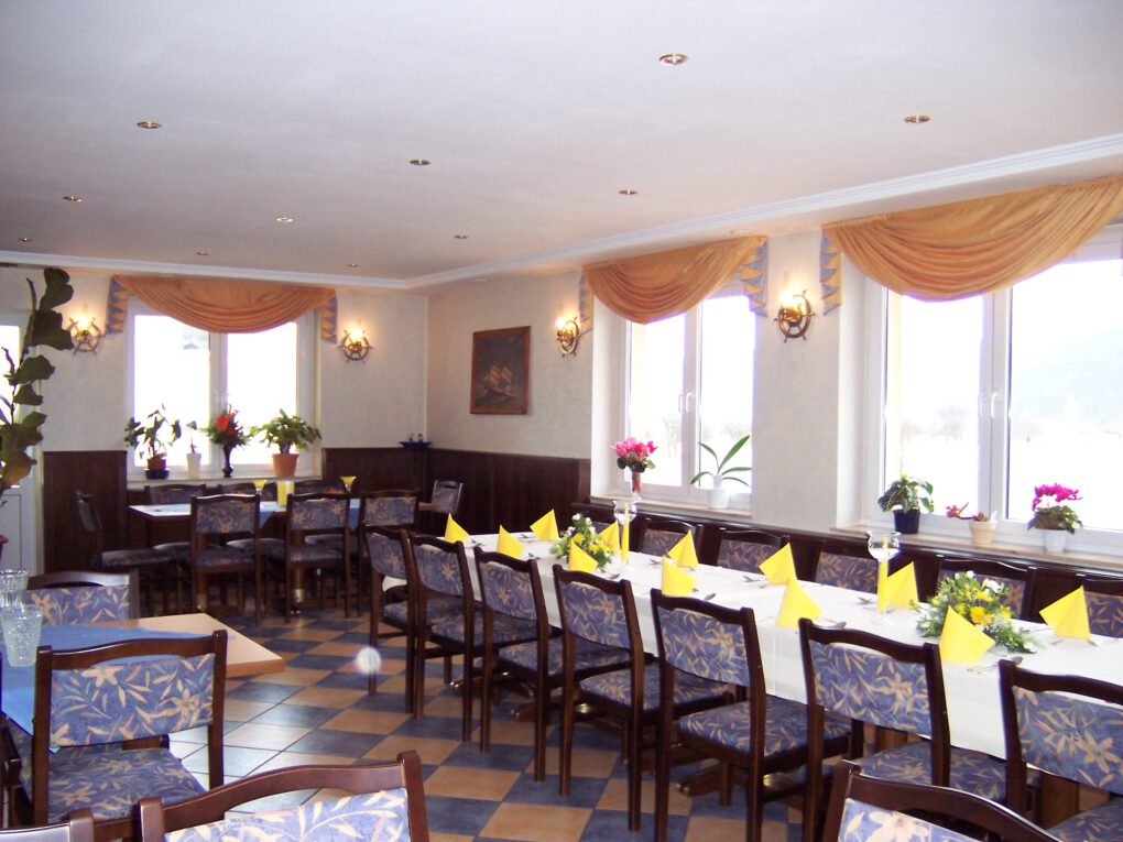 Ein heller, eleganter Speisesaal in Neumanns Dampfschiff Gaststätte ist mit langen Tischen mit gelben Servietten und Blumenschmuck ausgestattet. Große Fenster mit goldenen Vorhängen lassen natürliches Licht über blau gepolsterte Stühle herein.