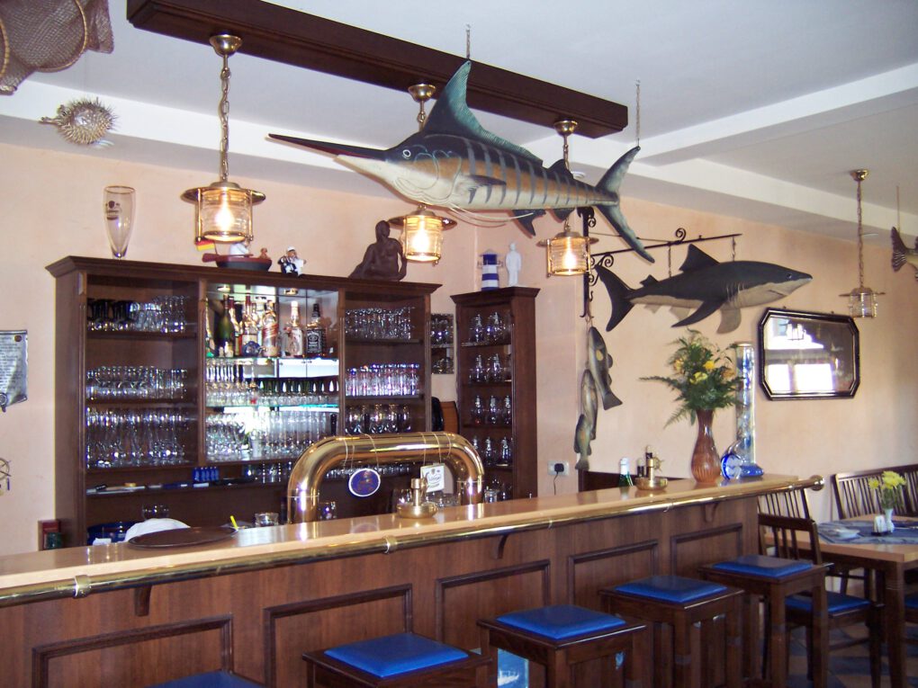 Die hölzerne Bar im Neumanns Dampfschiff Hotel ist mit Flaschen, Gläsern und nautischem Dekor ausgestattet, darunter ein Schwertfisch und ein Hai, die über dem Tresen angebracht sind. Blaue Barhocker säumen diese einladende Gaststätte.