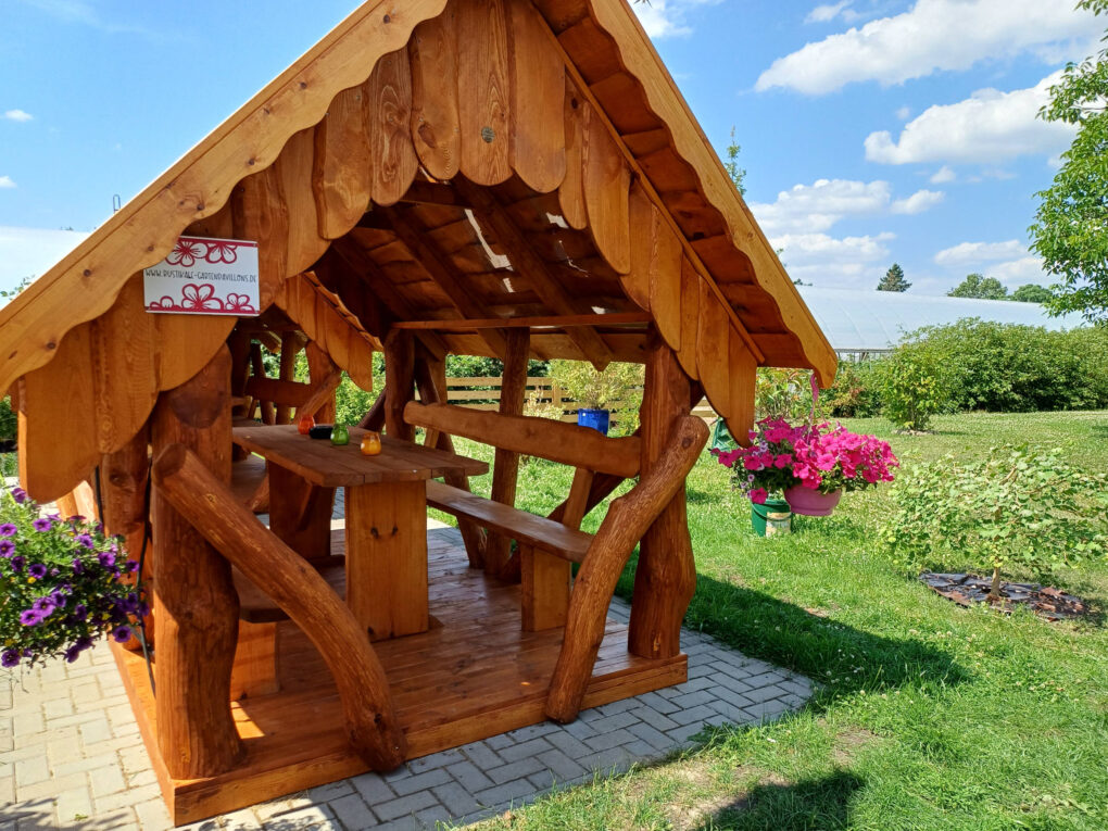 Ein rustikaler Holzpavillon mit Bänken und einem Tisch steht auf einer gepflasterten Fläche, umgeben von grünem Gras, Blumen und Pflanzen unter einem strahlend blauen Himmel im familiengeführten Hotel Sörnewitz.