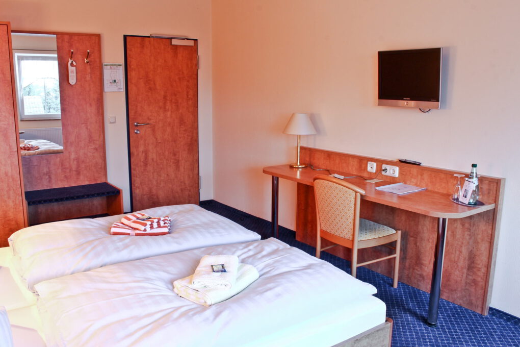 Ein ordentliches familiengeführtes Hotelzimmer in Sörnewitz mit zwei Einzelbetten, einem hölzernen Schreibtisch und Stuhl, einem an der Wand montierten Fernseher, einer Lampe, einer Flasche Wasser und Handtüchern, die ordentlich auf den Betten liegen. Im Hintergrund befinden sich eine Tür, ein Spiegel und ein Fenster.