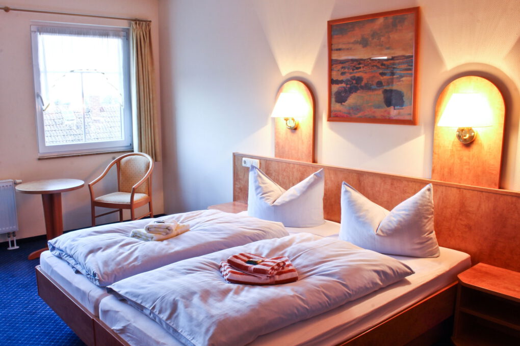 Ein aufgeräumtes Hotelzimmer in einem familiengeführten Hotel in Sörnewitz verfügt über zwei Einzelbetten mit weißer Bettwäsche, gefaltete Handtücher, Wandlampen, ein Bild über den Betten, ein Fenster sowie einen runden Tisch und zwei Stühle.