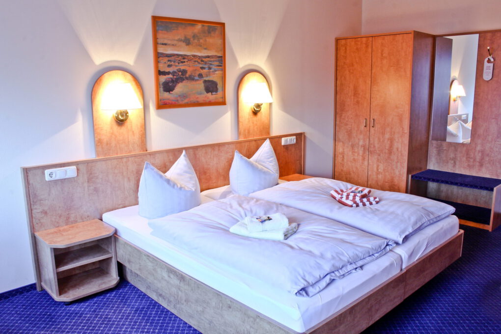 Ein ordentlich gemachtes Doppelbett mit weißer Bettwäsche, zwei Kissen und gefalteten Handtüchern steht in einem familiengeführten Hotel in Sörnewitz, mit Holzmöbeln, Wandlampen, einem Kleiderschrank, einem Spiegel und einem gerahmten Landschaftsgemälde über dem Kopfteil.