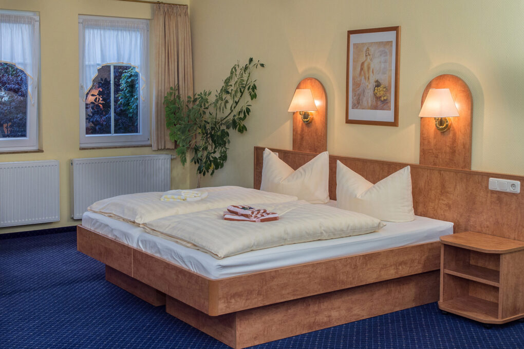 Ein ordentliches Zimmer im familiengeführten Hotel Sörnewitz verfügt über ein Doppelbett mit weißem Bettzeug, gefaltete Handtücher, zwei Wandlampen über dem Kopfende, ein gerahmtes Bild, eine Topfpflanze, einen blauen Teppich und zwei Fenster mit Vorhängen.